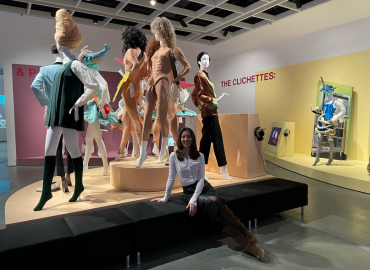 Woman in white top and black skirt with boots sitting on riser consisting of mannequins dressed in 1960s and 1970s outfits