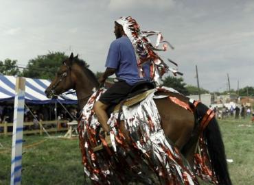 Still from Mohamed Bourouissa&amp;#039;s Horse Day. Young black man riding a brown horse decorated in red and silver ribbons.