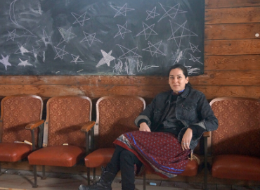 Crystal Migwans sitting at the end of a row of chairs in front of a blackboard with stars drawn on in chalk.